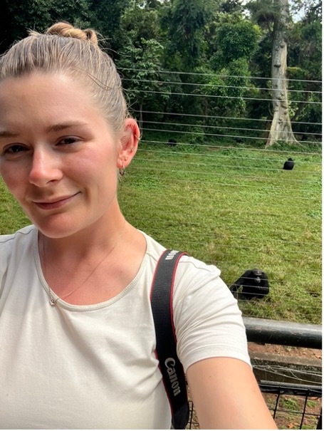 A woman taking a selfie outdoors with a grassy enclosure and chimpanzees visible behind a fence in the background.