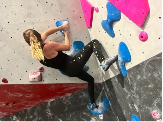 A woman rock climbing indoors on a wall with blue and pink holds, wearing athletic clothing and chalk on her hands.