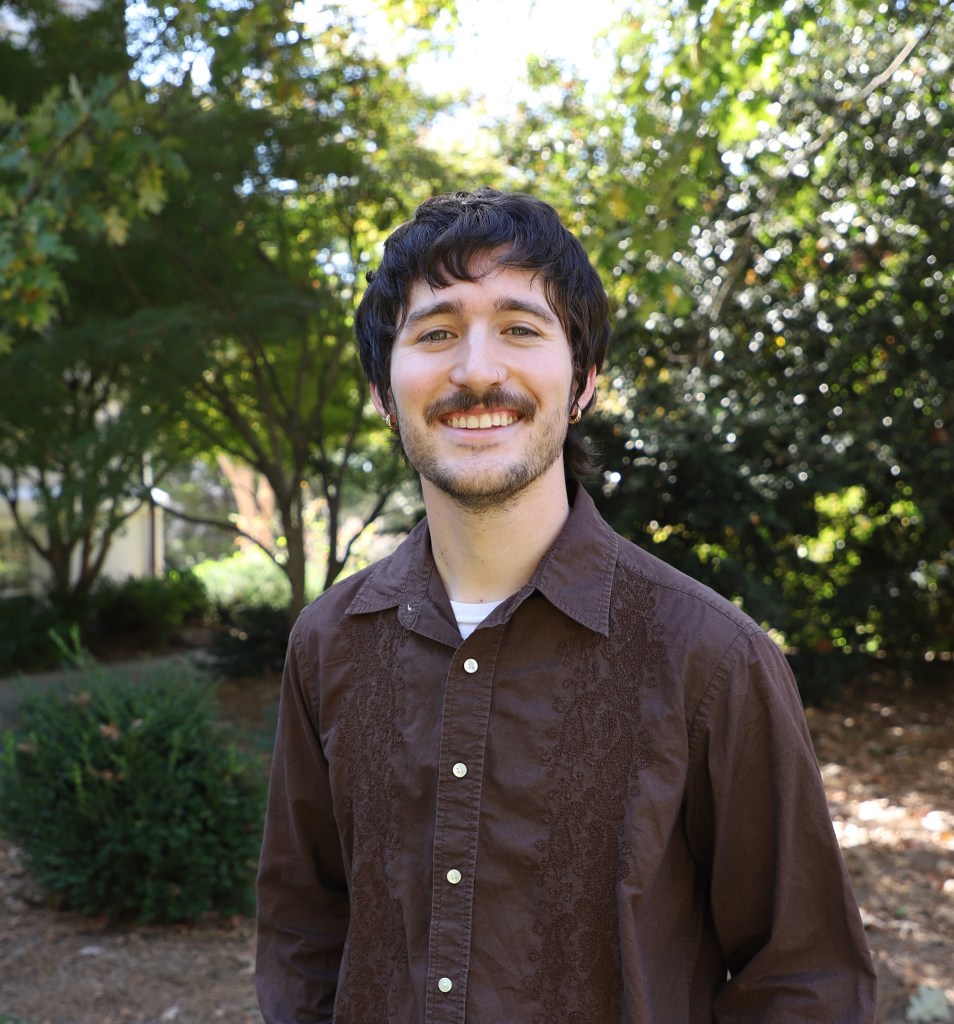 A smiling person with short dark hair and a mustache stands outdoors in a leafy park, wearing a brown button-up shirt, with trees and greenery softly blurred in the background.