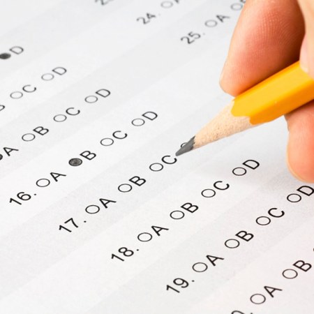 Close-up of a hand holding a sharpened yellow pencil over a multiple-choice answer sheet. Several bubbles are filled in next to numbered questions, and the pencil tip is positioned near one of the answer options.
