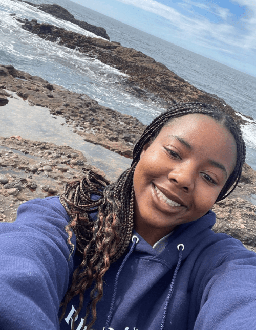 A smiling young woman with braided hair is taking a selfie in front of rocky shoreline and ocean.