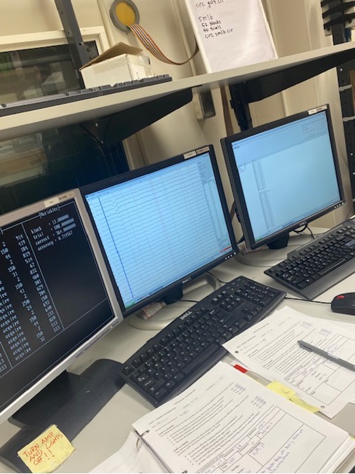 A workspace featuring three computer monitors displaying various data, with a keyboard and paperwork on the desk.